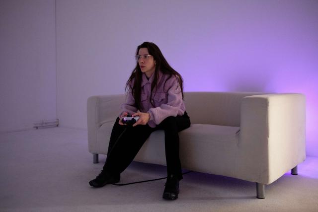 Porträt der multidisziplinären Künstlerin Mary-Audrey Ramirez, die ihr Spiel Forced Amnesia spielt. Sie sitzt auf einem Sofa, in violettes Licht getaucht, und hält einen Videospiel-Controller in der Hand.