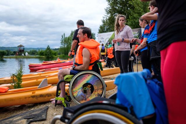 Homme dans un fauteuil roulant au bord d'un lac. Il porte une veste de secours et on voit des kayaks derrière lui.