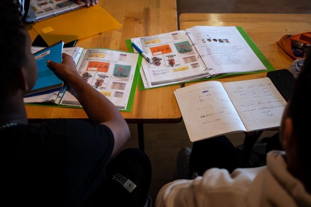 Elèves dans une salle de classe, on les voit de dos avec des livres de classe ouverts sur une table.