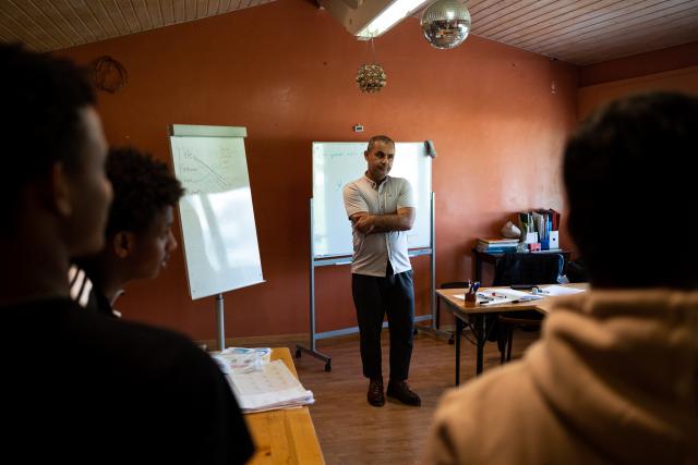 Elèves dans une salle de classe, on les voit de dos et l'enseignant de face devant deux tableaux.