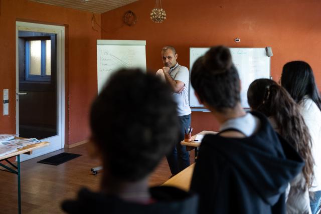 Schüler*innen in einem Klassenzimmer, von hinten gesehen, mit der Lehrkraft, die ihnen gegenüber vor zwei Tafeln steht.