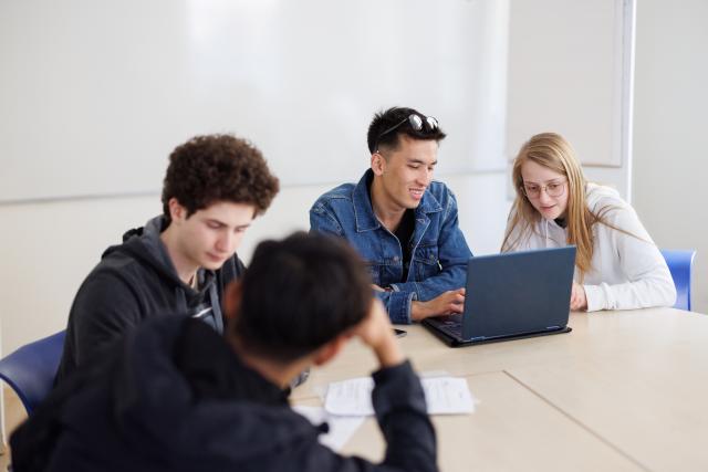 4 students sitting around a table. Two of them are looking at something on a laptop.