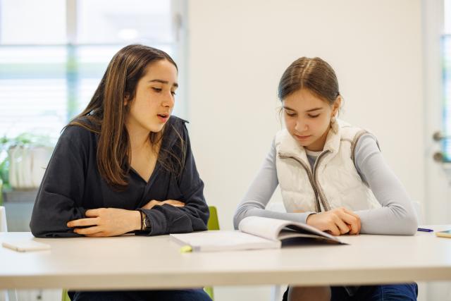 Two girls sitting at a table reading a book.