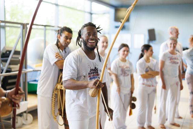 portrait de Bruno Jordan Lucio de Oliveira du Capoeira Team Luxembourg asbl. Il est entouré d'autres personnes et tient un instrument brésilien entre ses mains.