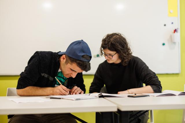 Two students sitting at a table. One of them is writing in a notebook.