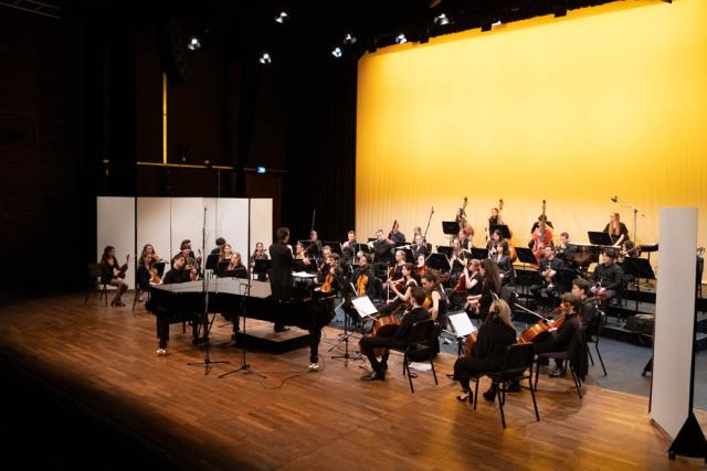 Musiker*innen eines Orchesters auf einer Bühne, die in Gelb beleuchtet ist.