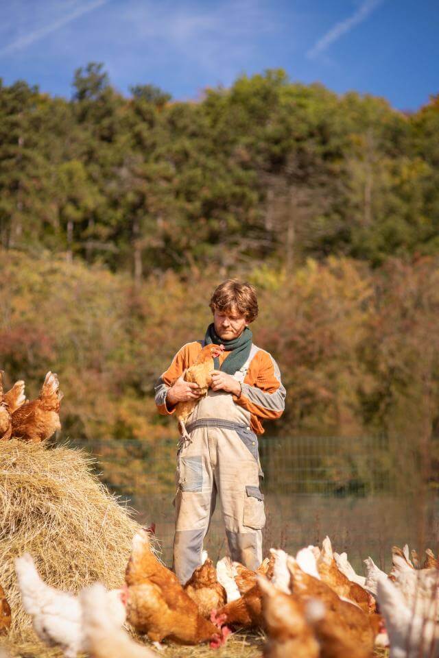 Tom Kass encerclé de poules. Il en tient une dans son bras.